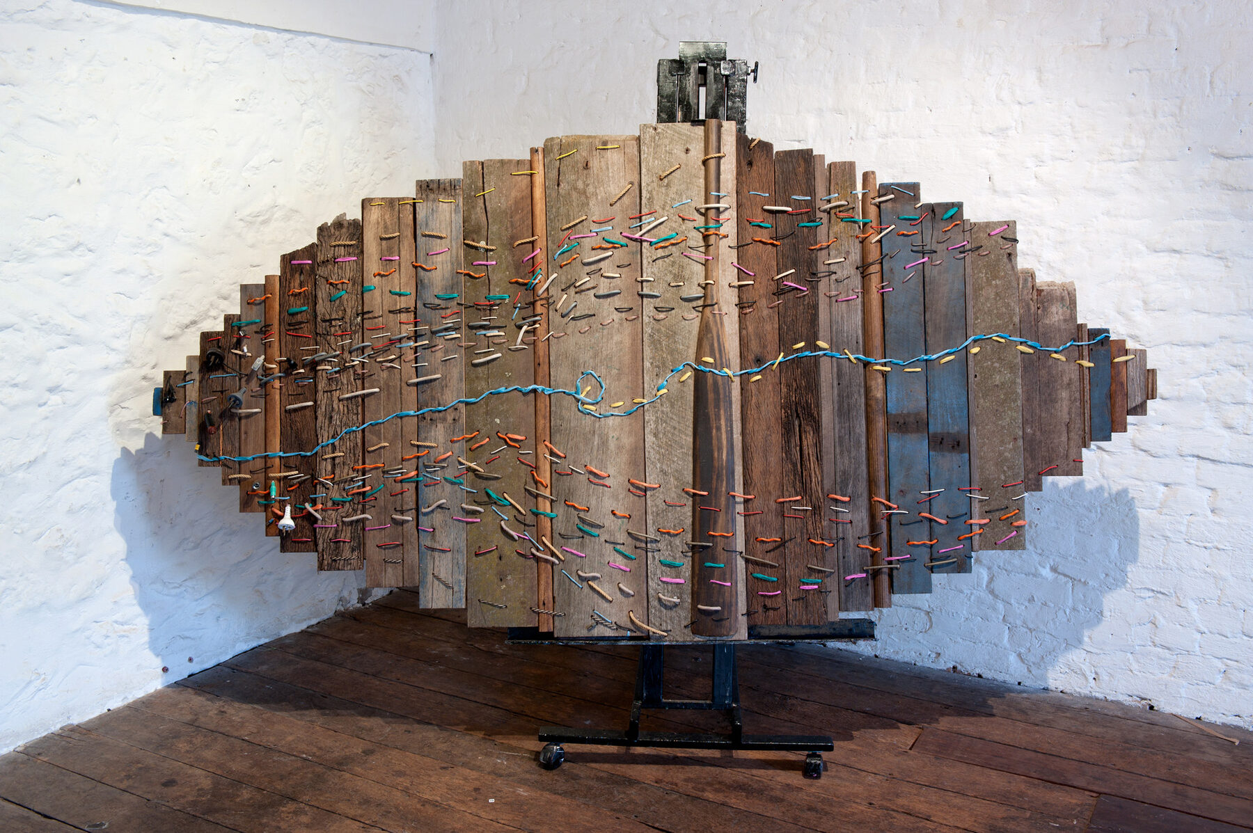 A large lens-shaped assemblage of weathered vertical timber boards, stitched across with blue and coloured thread, standing in a white-walled gallery.