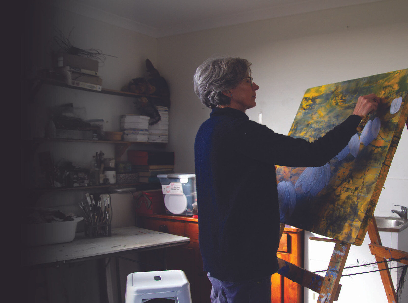 Catherine Higham in her studio, standing at an easel and working on a painting in yellows, greens and pale blue, with shelves of materials behind her.