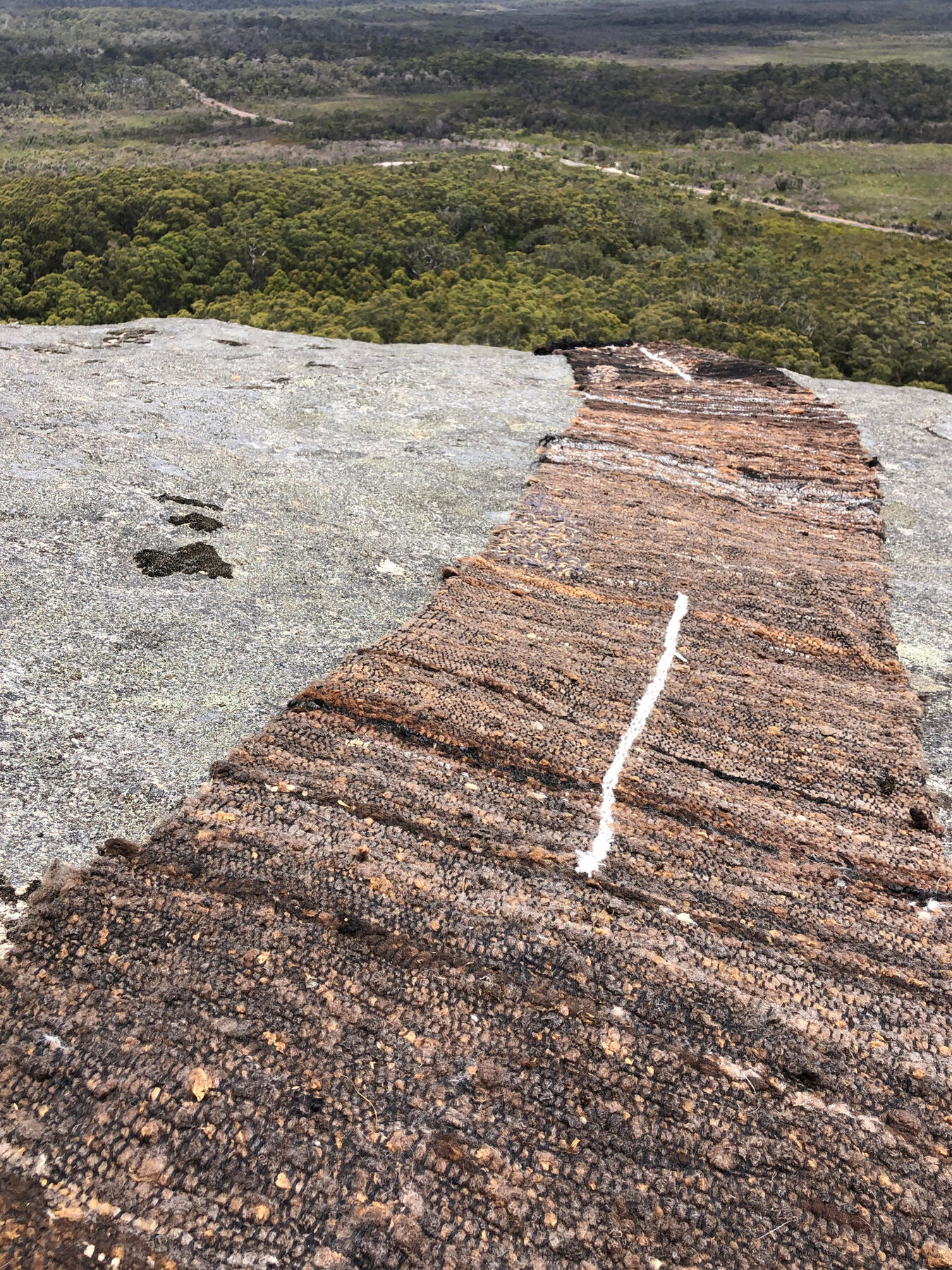 A long runner of brown felted wool and alpaca fibre laid across a pale granite outcrop, with bushland and distant hills beyond.